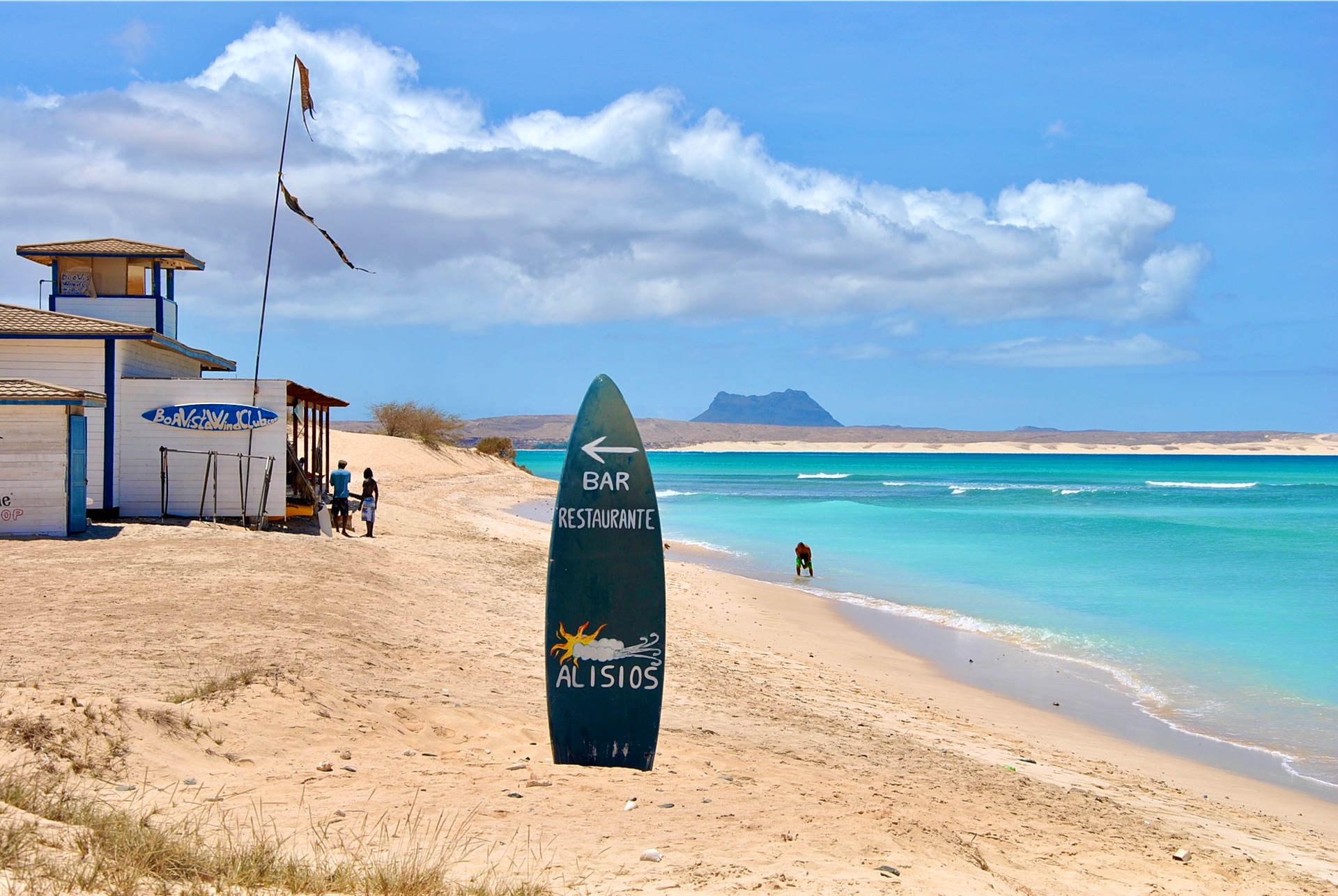 Tavola da surf con l'insegna di un bar sulla spiaggia, mare turchese e una montagna all'orizzonte, a Boa Vista