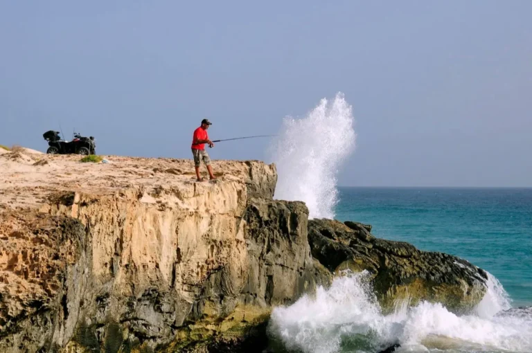 Pescatore in rosso sulla scogliera mentre un'onda esplode sotto di lui